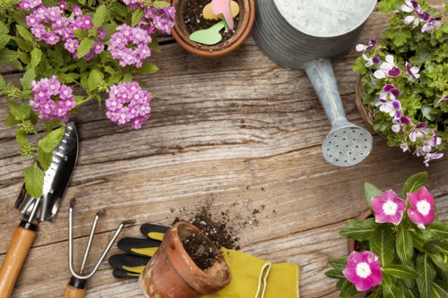The image shows a rustic wooden outdoor table set in a garden area, with a weathered surface and natural wood grain visible. Several potted flowering plants with purple, pink, and white blossoms are arranged on and around the table, adding vibrant colour and a natural aesthetic typical of a well-maintained garden. Garden tools, including a trowel with a wooden handle, a small hand rake, a yellow gardening glove, and a terracotta pot with soil spilling out, are placed on the table, indicating ongoing planting or potting activities. A metal watering can with a long spout is also present, suggesting water is being prepared for the plants. The scene captures a calm outdoor environment, likely part of a residential garden, with natural light illuminating the setup, emphasizing the textures of the soil, plant leaves, and wooden surface. The overall arrangement reflects a gardening scene where plants are being cared for or prepared for planting, aligning with services such as hedge trimming and garden maintenance provided by Hedge Trimming Covent Garden.