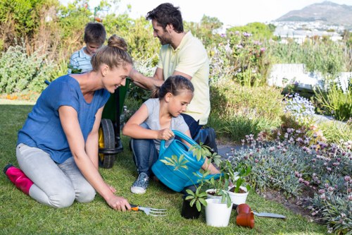 A family of four engaged in gardening activities in a well-maintained outdoor garden space. The scene features a lush green lawn bordered by flower beds with a variety of blooming plants and shrubs. In the foreground, a woman wearing a blue t-shirt and grey trousers is kneeling on the grass, using a small gardening fork to tend to a young plant. Beside her, a girl dressed in a blue top and jeans is sitting on a gardening stool, holding a blue watering can with a plant inside, preparing to water the plants. In the background, a man in a light-colored shirt and beige trousers is using a pair of scissors or clippers to trim a hedge or shrub, while another child, dressed in a striped hoodie, observes nearby. The garden is bathed in natural daylight under a partly cloudy sky, with a scenic view of distant hills and a residential area visible in the background. The scene highlights outdoor family gardening under mild weather conditions, with attention to detailed plant care, suitable for professional gardening or hedge trimming services in Covent Garden or nearby areas.