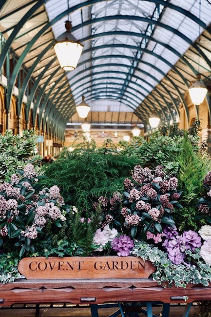 Inside a glass greenhouse with a curved, metal and glass arched ceiling illuminated by hanging lantern-style lights, a lush arrangement of various plants and flowers is displayed. The display includes