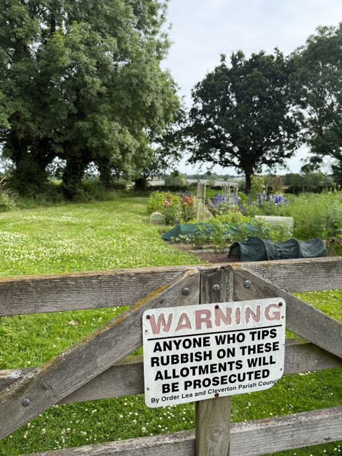 A rustic wooden garden gate with a warning sign attached, reading 'WARNING: Anyone who tips rubbish on these allotments will be prosecuted by Clevendon Parish Council and Order Lea.' Behind the gate, 